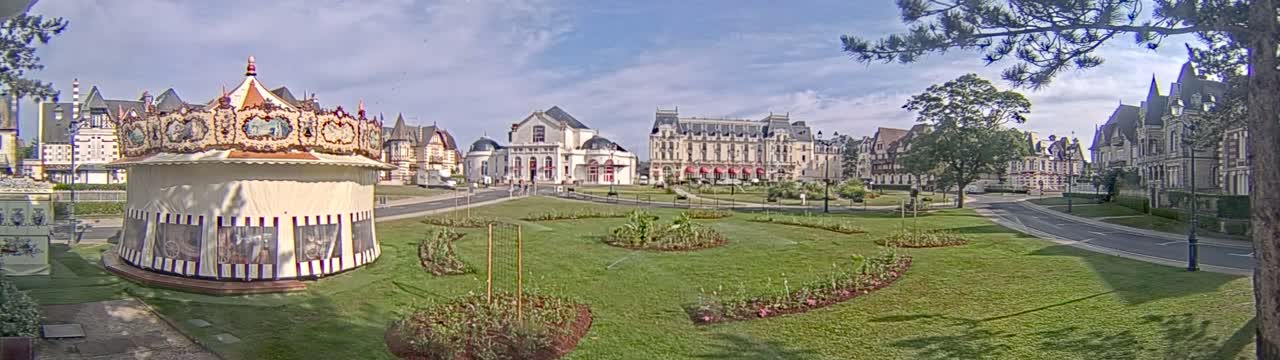 A carousel sits in a grassy town square surrounded by ornate buildings under a partly cloudy sky.