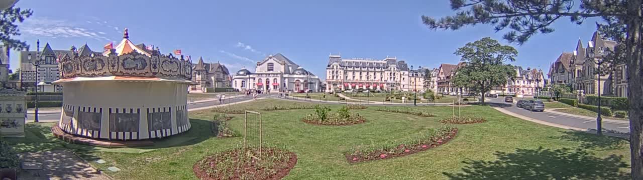 A carousel sits in a grassy town square in front of several large buildings, under a sunny sky.