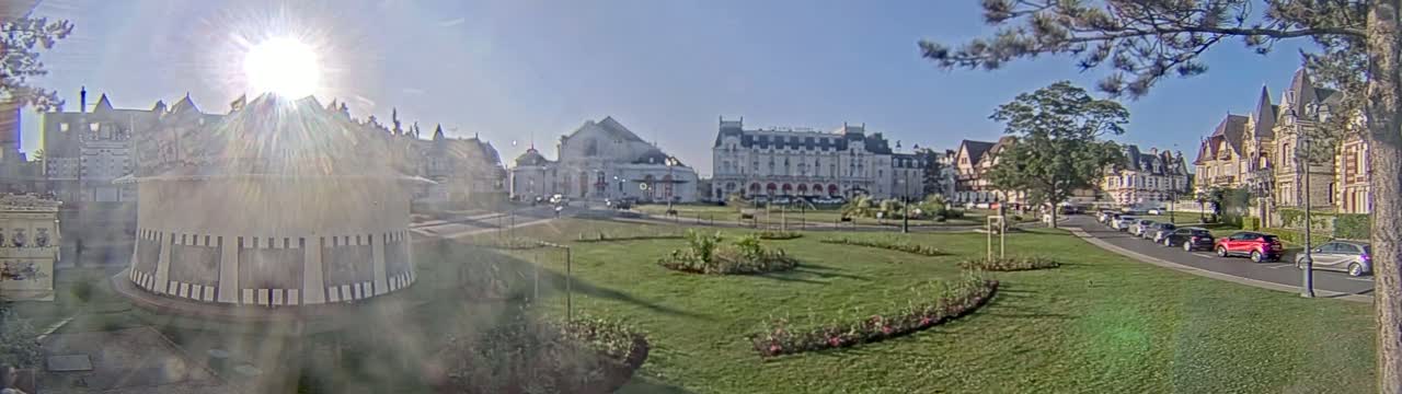 A sunny day reveals a plaza with several European-style buildings, manicured lawns, and parked cars.