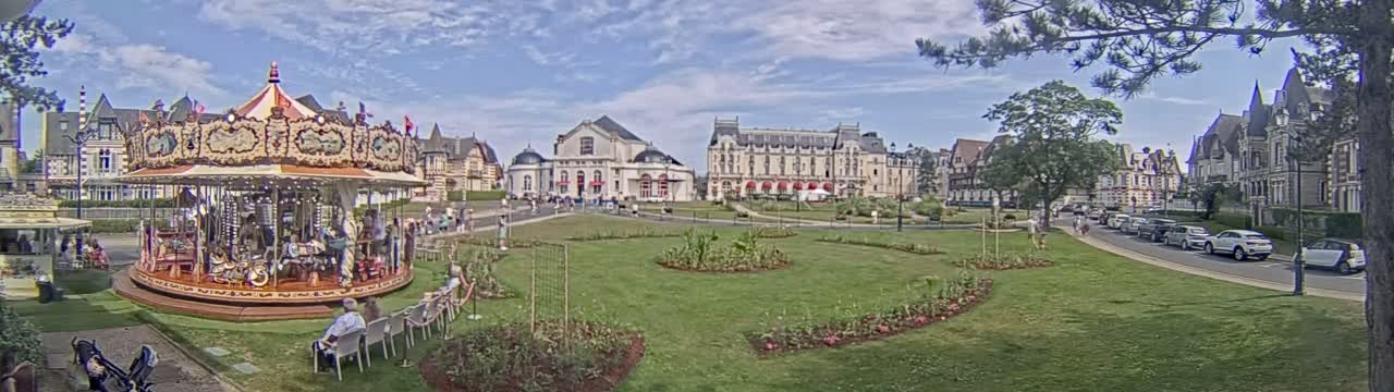 A carousel sits in a grassy town square next to several large buildings, under a partly cloudy sky.