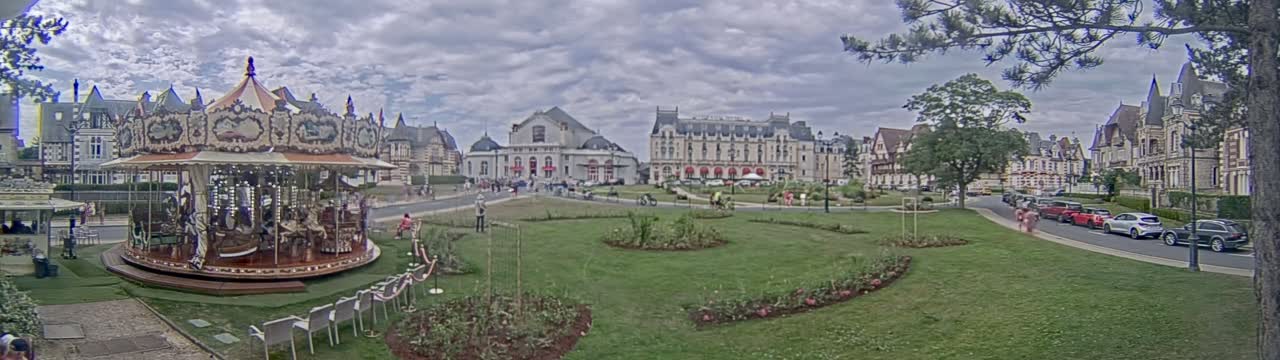A carousel sits in a grassy town square with several buildings behind it, under a cloudy sky.