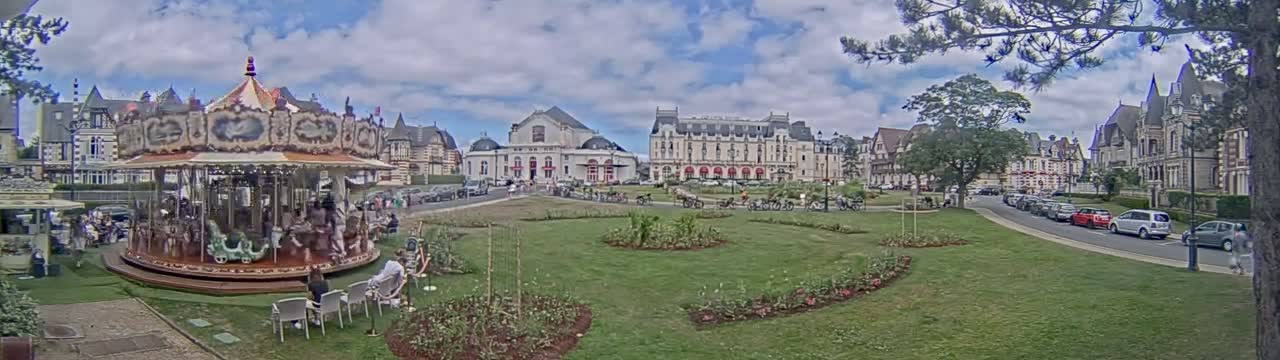 A carousel sits in a grassy town square under a partly cloudy sky, with several buildings and parked cars visible in the background.