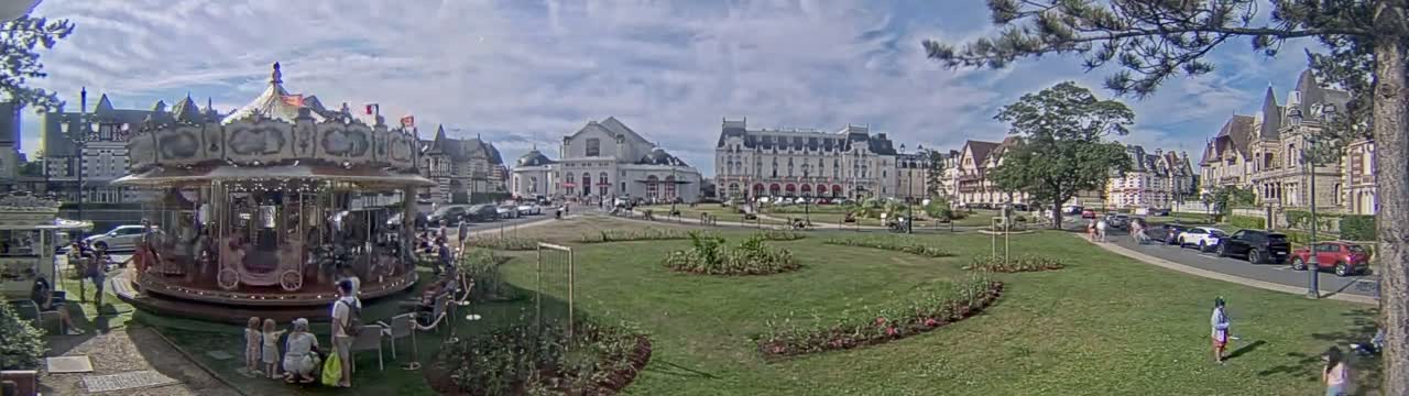 A carousel sits in a grassy town square in front of several large buildings under a partly cloudy sky.
