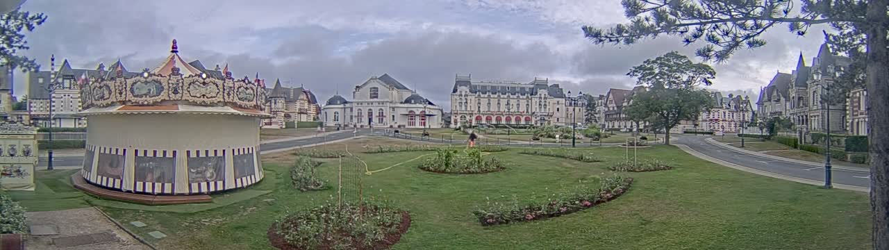 A carousel sits in a grassy park, overlooking a street and several ornate buildings under a cloudy sky.