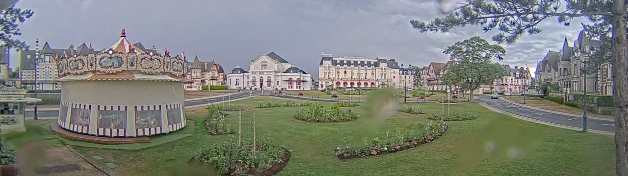 A carousel sits in a grassy park across from several large buildings under an overcast sky.
