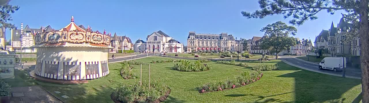 A carousel sits in a grassy park in front of several large buildings on a sunny day.