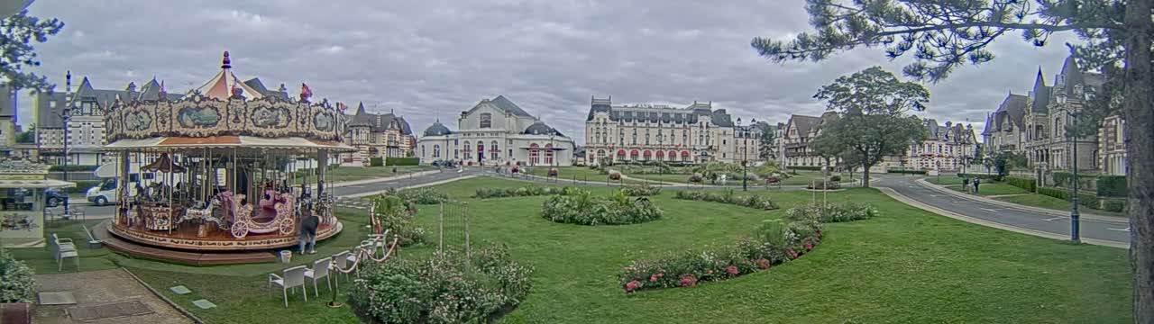 A carousel sits in a grassy town square, surrounded by several large buildings under an overcast sky.