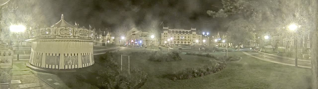 A carousel sits in a grassy town square, surrounded by several large buildings under an overcast sky.