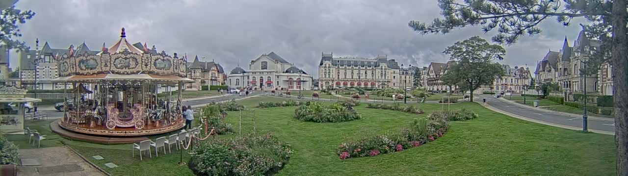 A carousel sits in a grassy town square under an overcast sky, surrounded by several large buildings.
