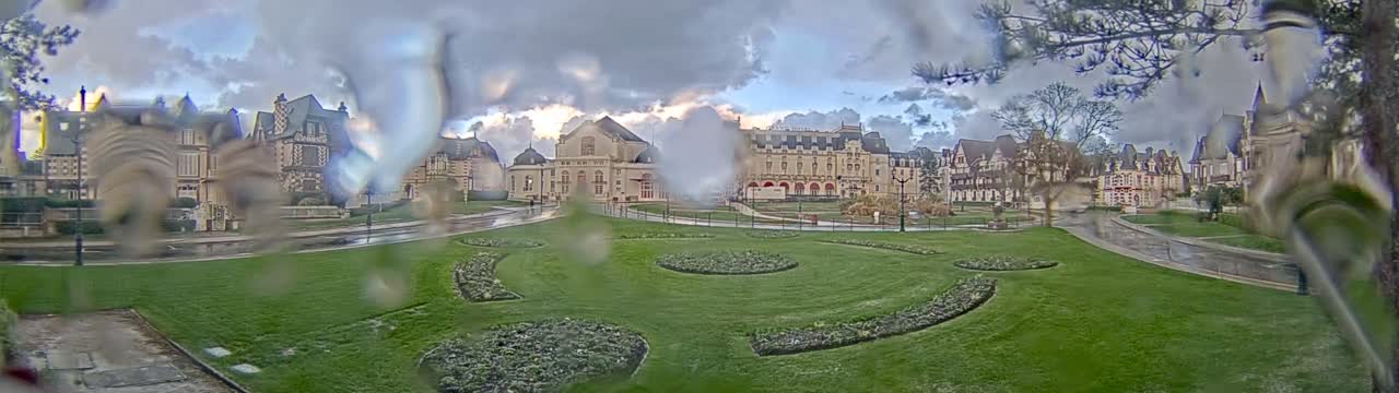 Stately, ornate buildings of European architecture surround a large, green park with manicured lawns and decorative garden beds, viewed through a rain-splattered lens under a cloudy sky with glimpses of light.