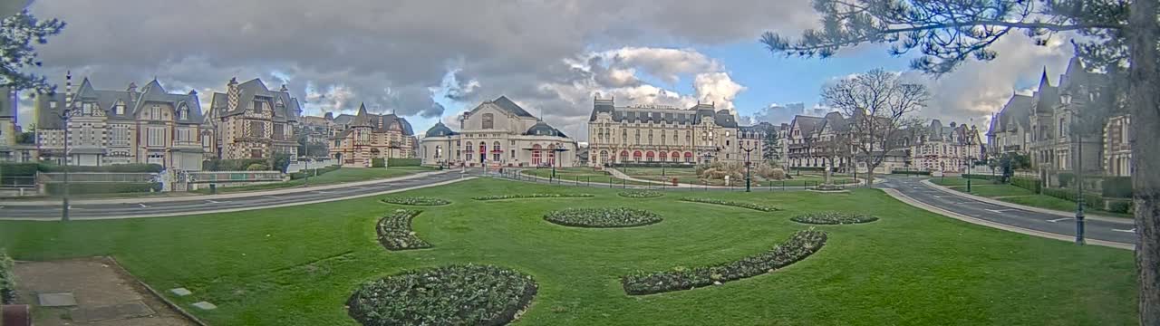 A panoramic view captures a meticulously landscaped urban park with green lawns and decorative flower beds, bordered by curved roads and an array of elaborate architectural buildings including a grand hotel and a domed theater, all beneath a partly cloudy sky.