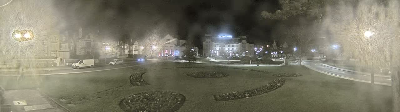 A panoramic view captures a meticulously landscaped urban park with green lawns and decorative flower beds, bordered by curved roads and an array of elaborate architectural buildings including a grand hotel and a domed theater, all beneath a partly cloudy sky.