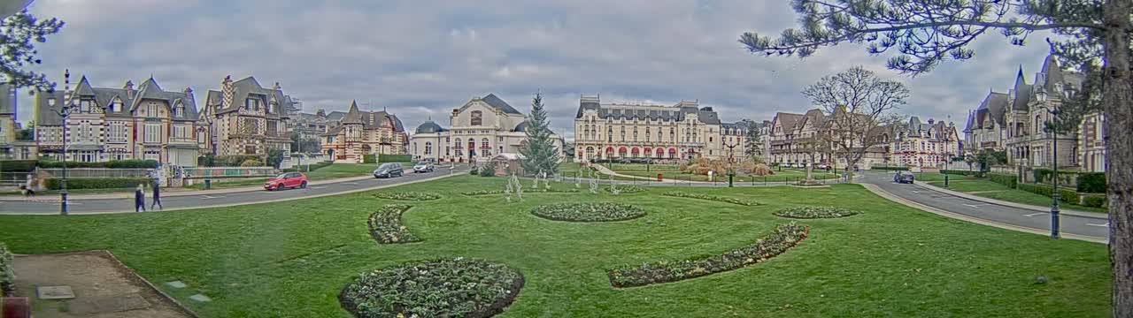 A panoramic view captures a meticulously landscaped urban park with green lawns and decorative flower beds, bordered by curved roads and an array of elaborate architectural buildings including a grand hotel and a domed theater, all beneath a partly cloudy sky.