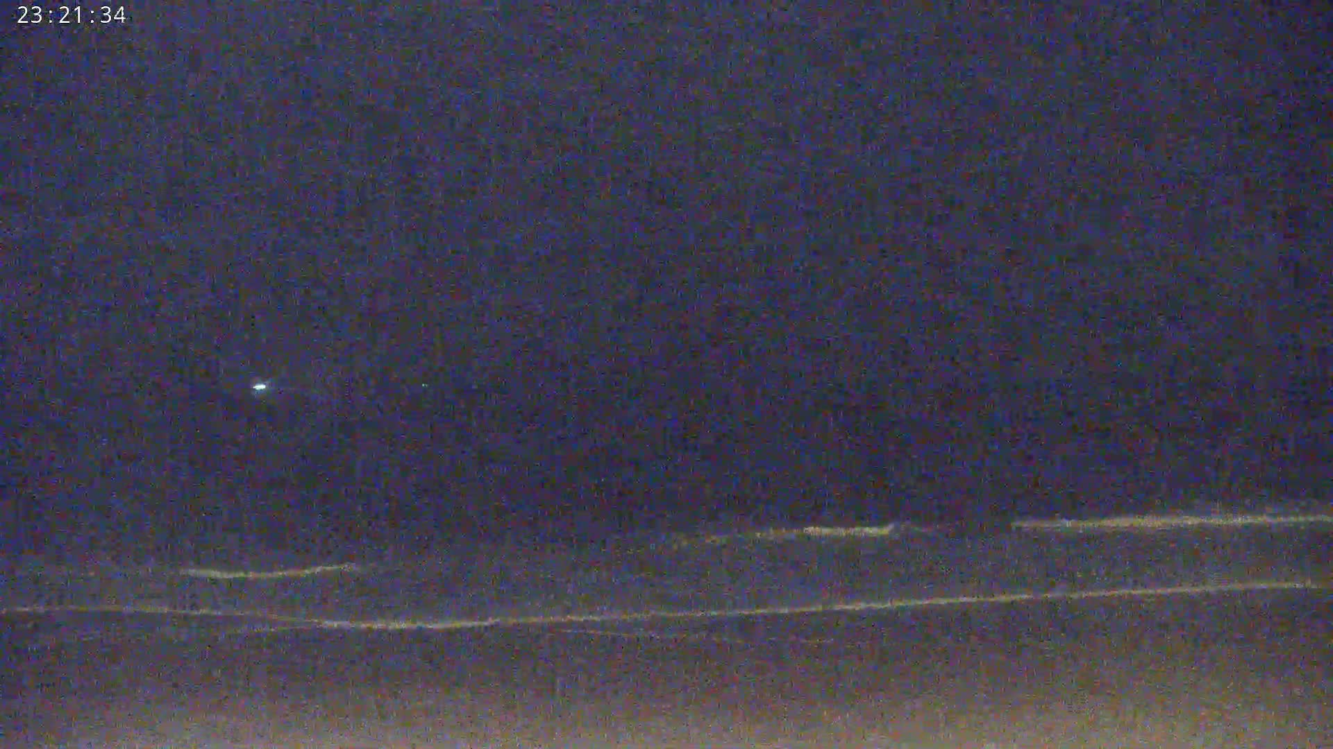 A nighttime beach scene shows dark water with small waves lapping the shore under a dark sky with a single, bright light visible.