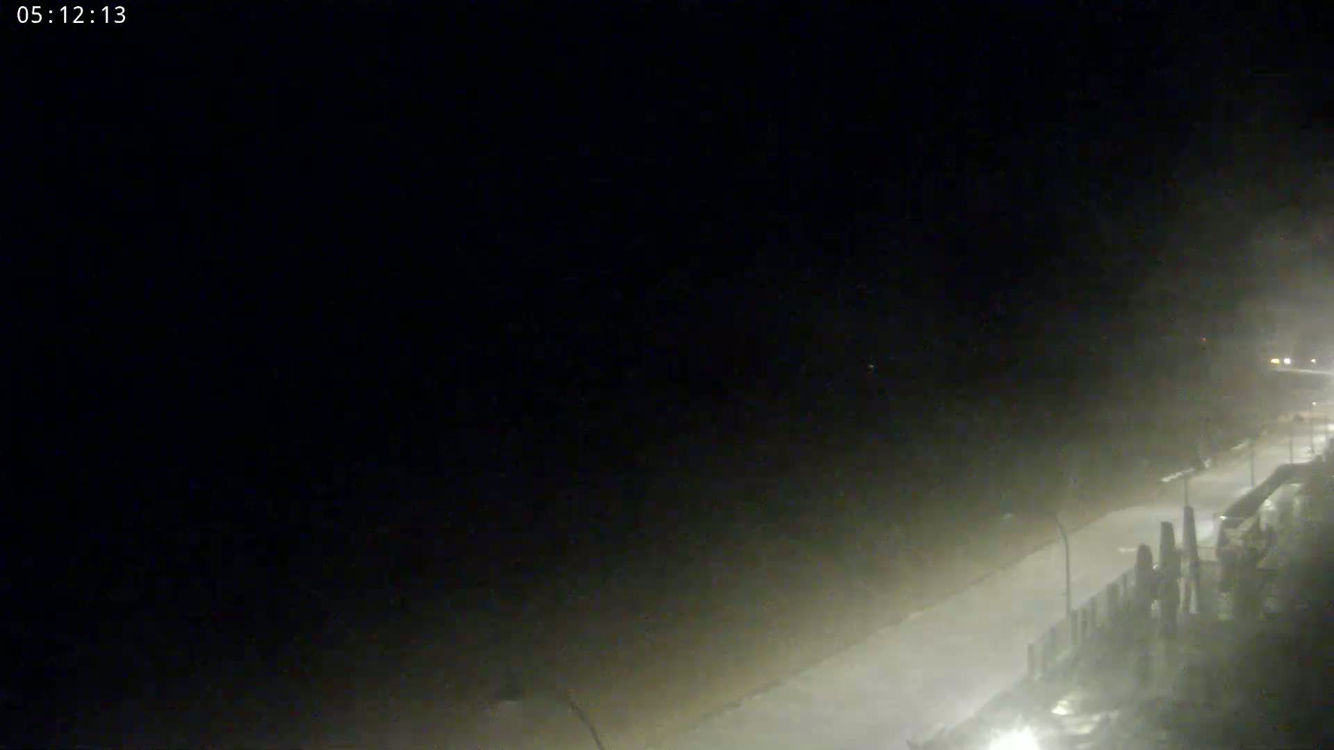A nighttime, hazy view shows a dimly lit road next to a structure, possibly a seawall, under heavy fog or mist.