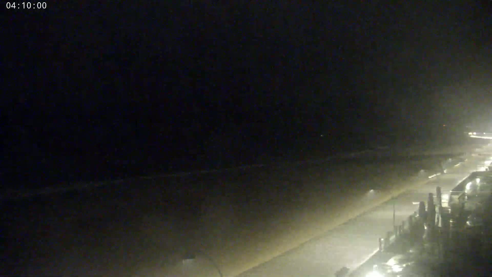 A nighttime view of a dimly lit beach and road, with faint waves visible in the distance under a dark, overcast sky.