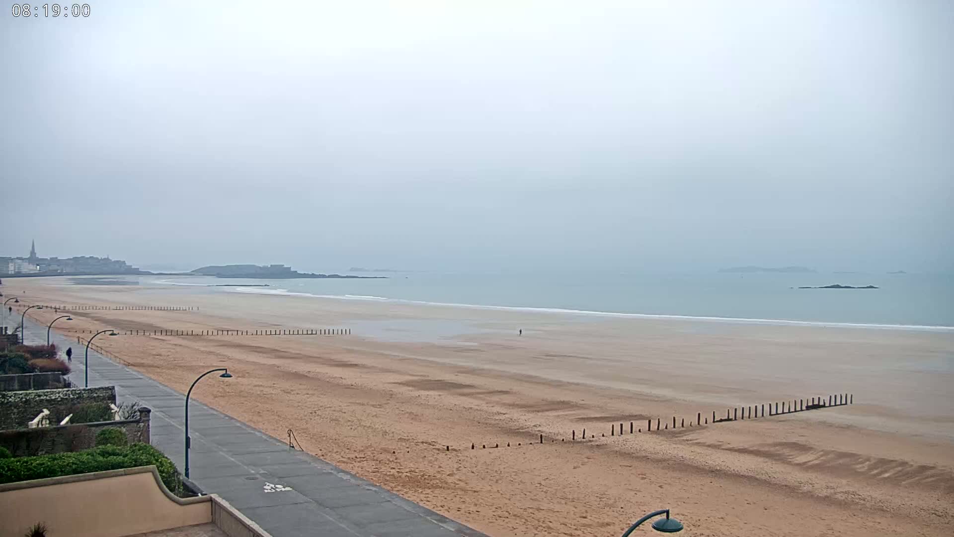 A wide sandy beach stretches to a calm, hazy ocean under an overcast sky, with a paved walkway and distant town visible.