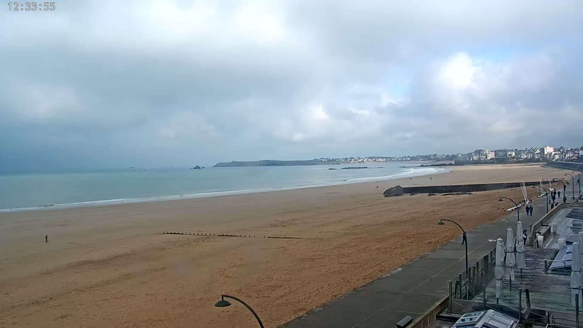 A wide, sandy beach stretches out to a calm sea under a cloudy sky, with a town visible in the distance.