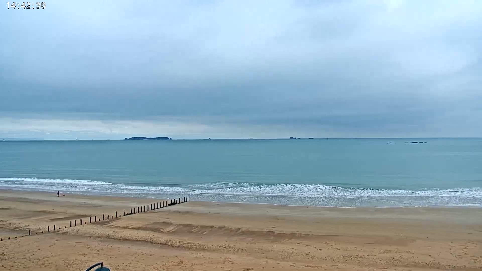 A sandy beach with gentle waves under a cloudy sky stretches to a calm ocean with distant islands.
