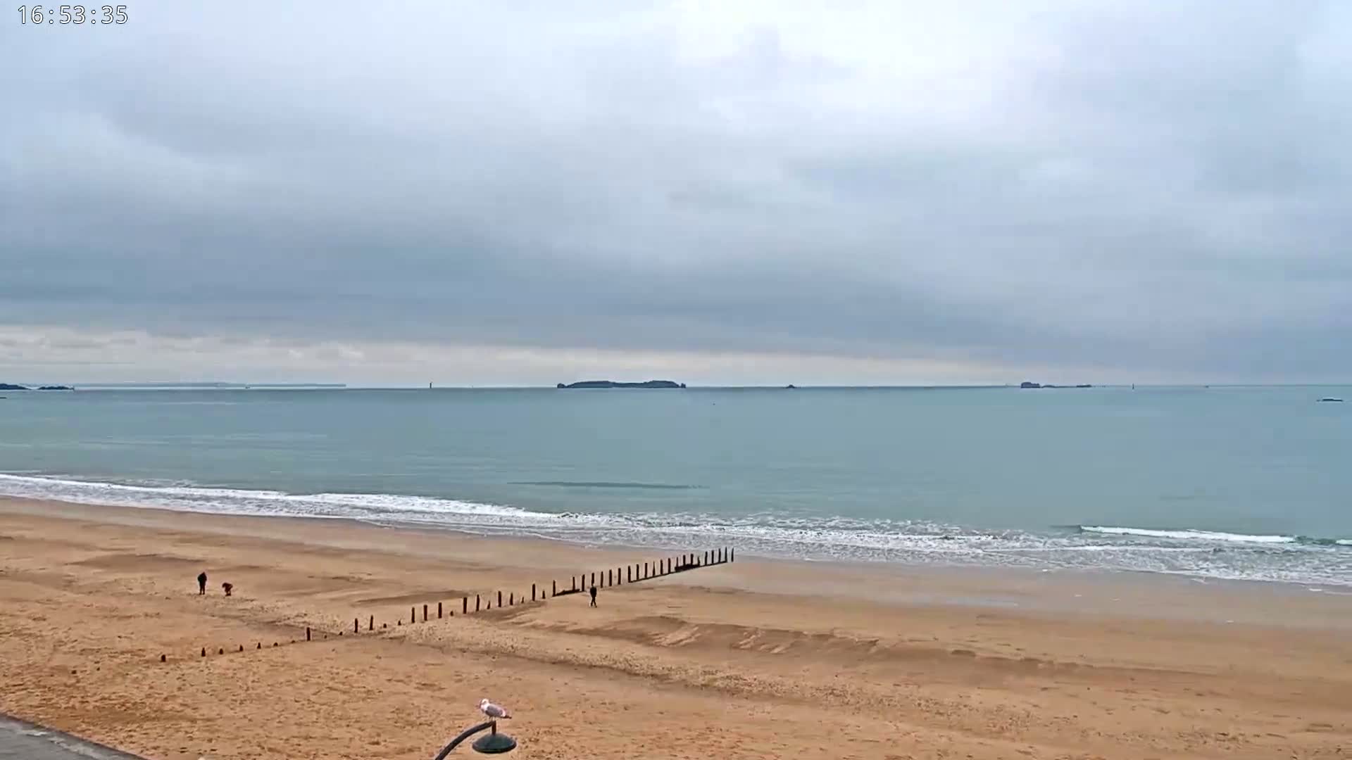 A sandy beach with a calm sea under an overcast sky, showing small waves lapping the shore, and a few people walking near a line of wooden posts.