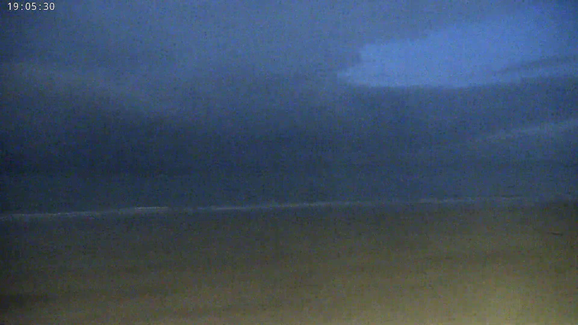 A dark, overcast sky hangs over a calm, flat body of water and a dark beach at twilight.