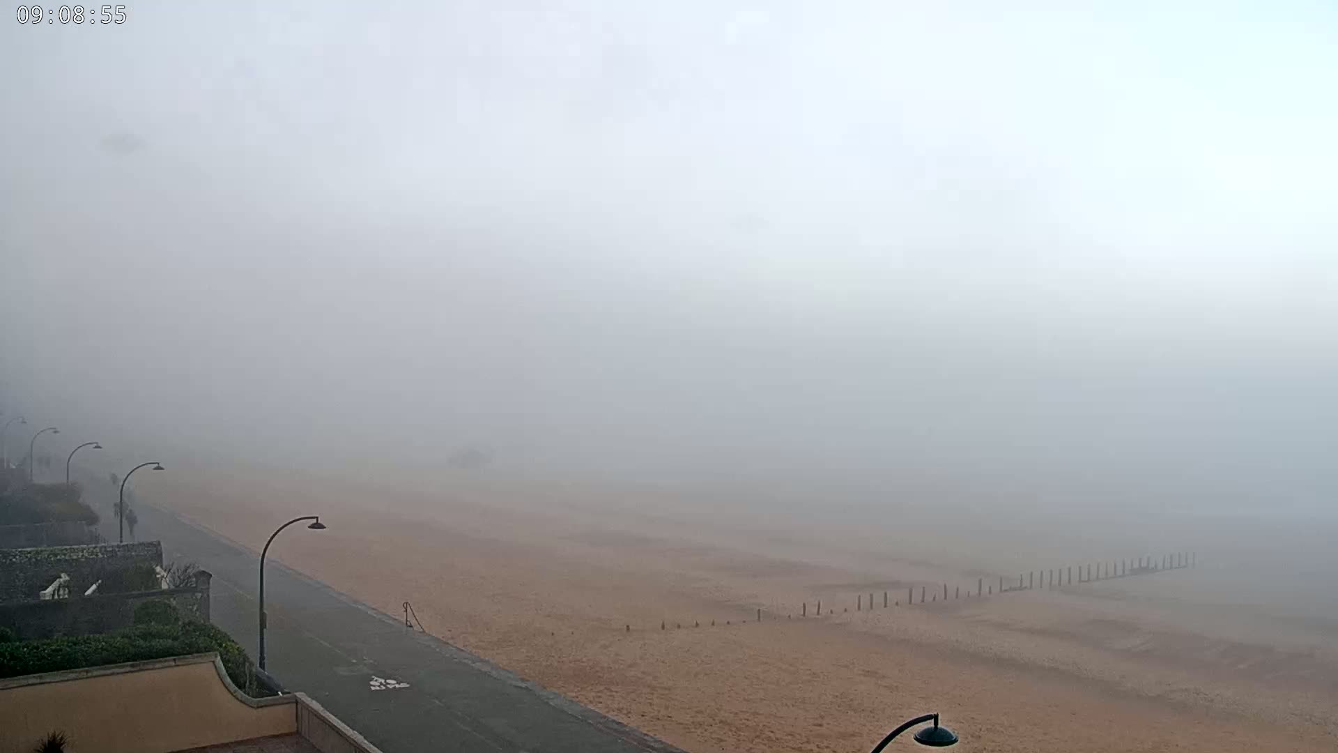 A foggy beach scene is visible from a high vantage point, showing a sandy beach, a paved walkway with streetlights, and some buildings in the distance.