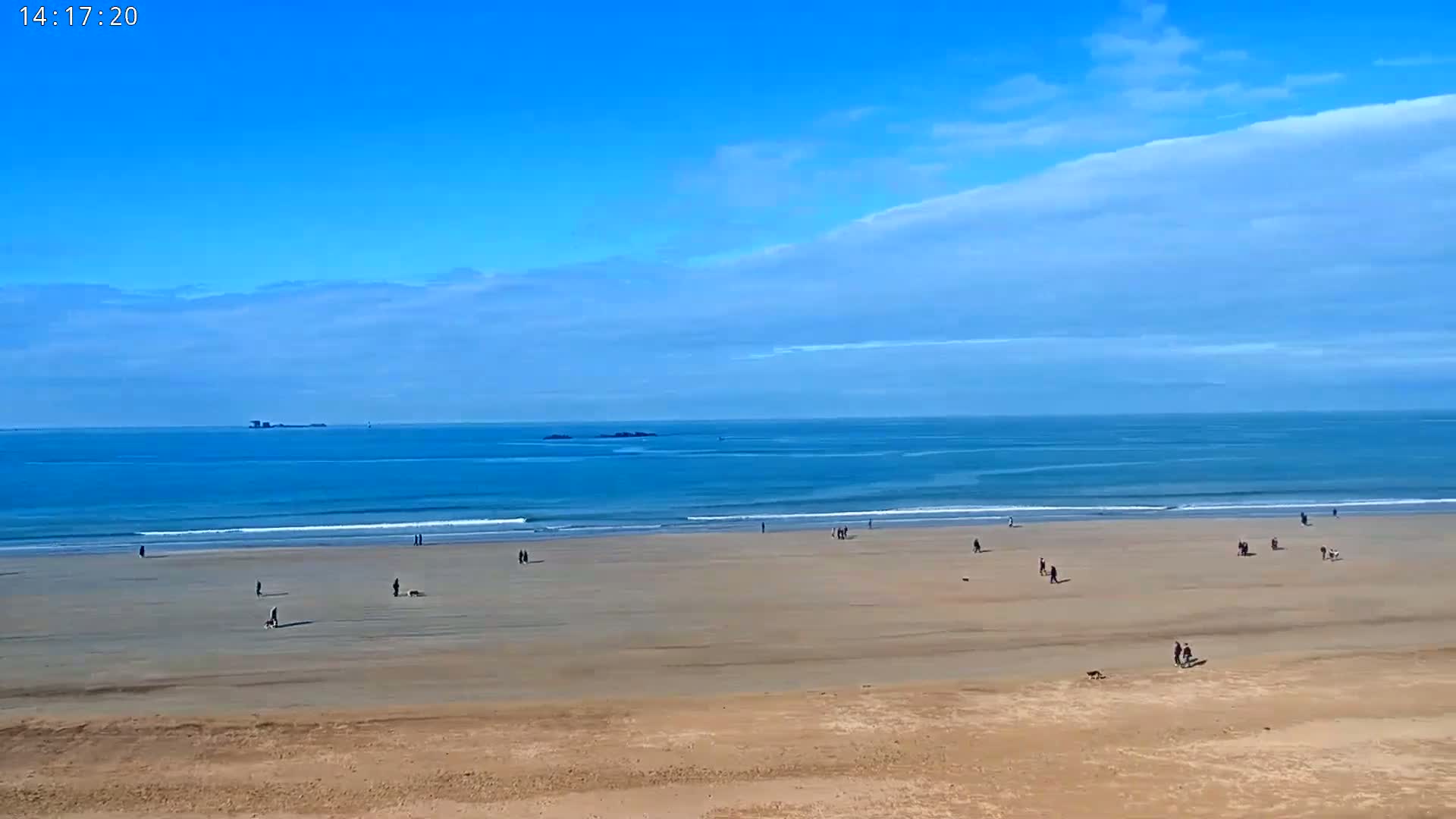 A wide sandy beach stretches to a calm blue ocean under a mostly clear blue sky with some wispy clouds.