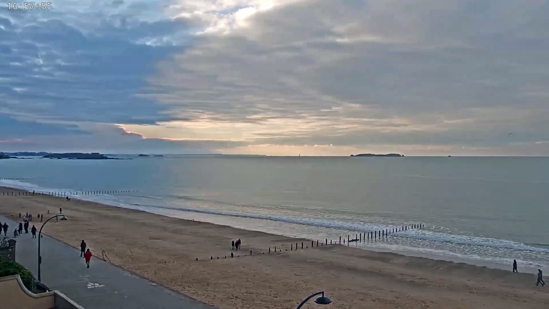 A mostly cloudy sky overlooks a sandy beach with calm waves and several people walking along the shore and a paved walkway.
