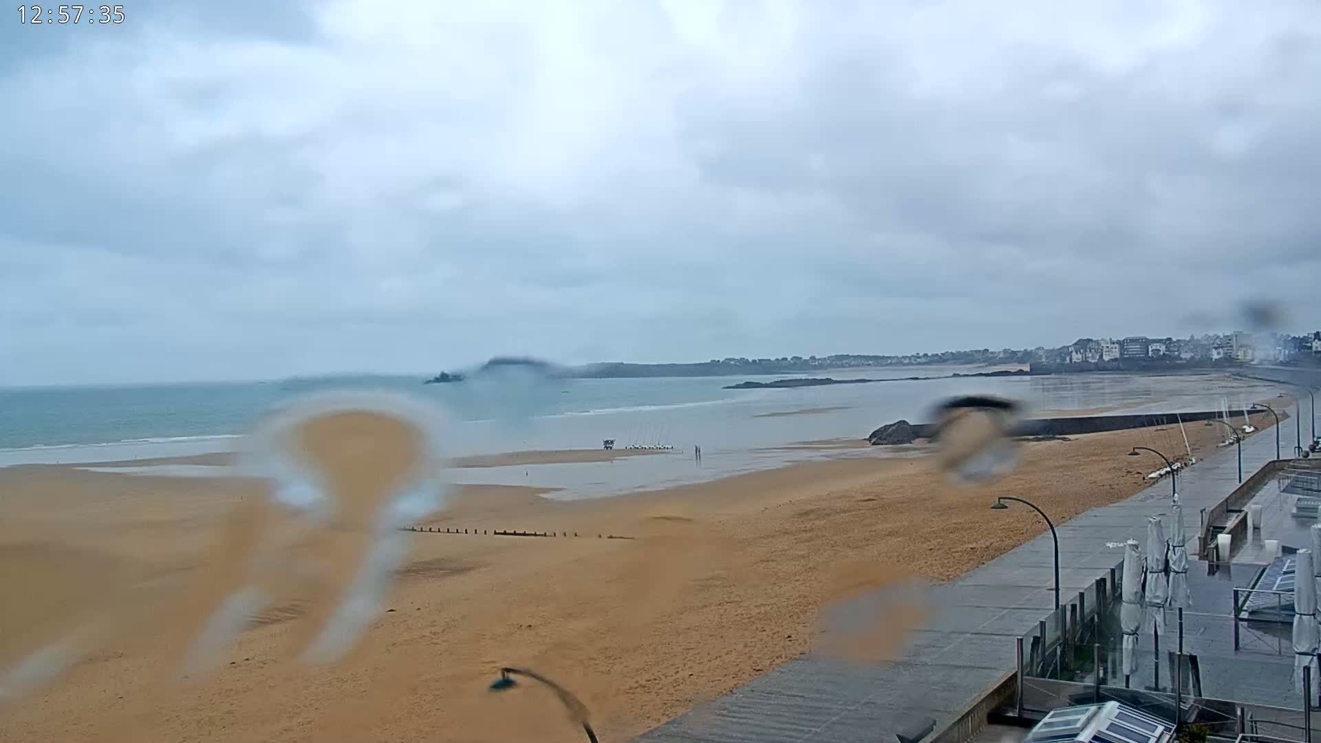 A rain-covered window shows a view of a wide sandy beach, calm ocean, and a distant town under a cloudy sky.