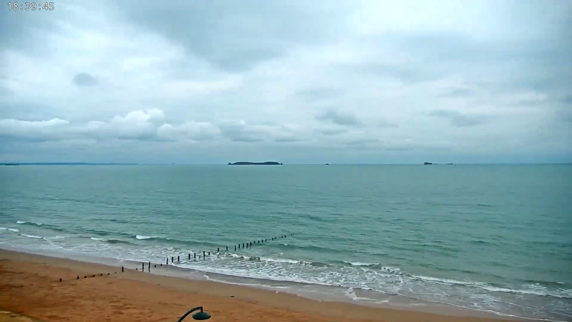 A sandy beach with gentle waves rolls onto the shore under a mostly cloudy sky, with islands visible in the distance.