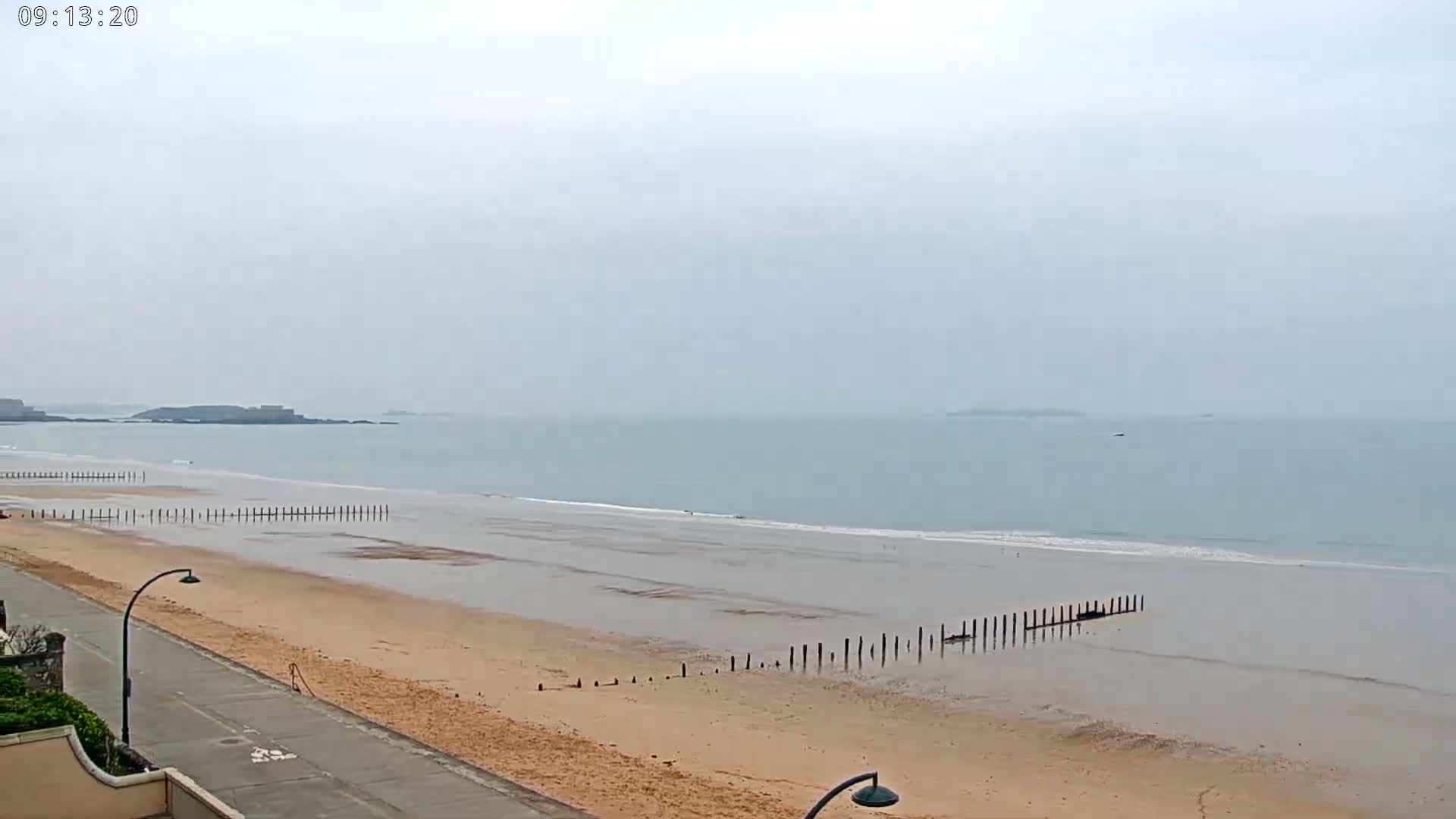 A sandy beach stretches out to a calm, light-blue ocean under an overcast sky, with dark wooden posts extending into the water.
