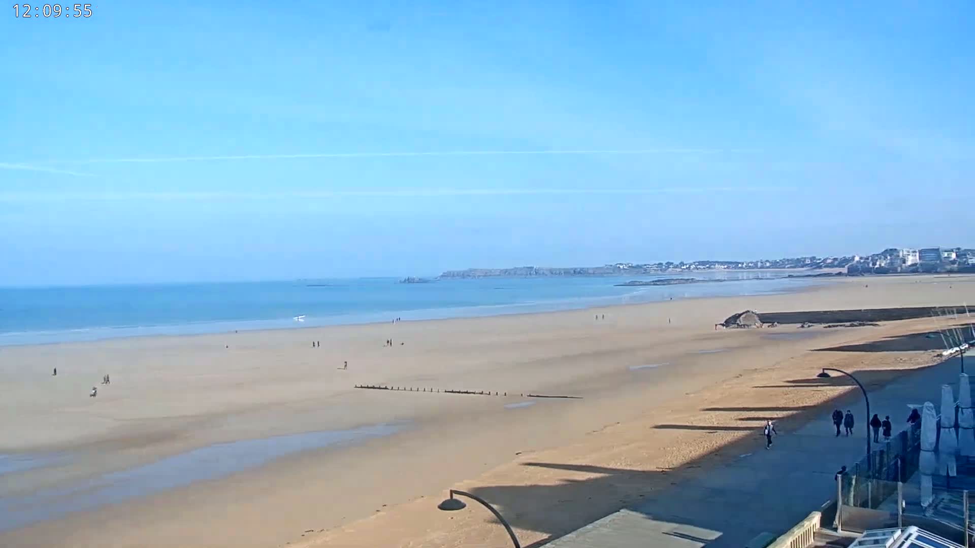 A wide, sandy beach stretches to a calm ocean under a clear blue sky, with a few people walking along the shore and on the sand.