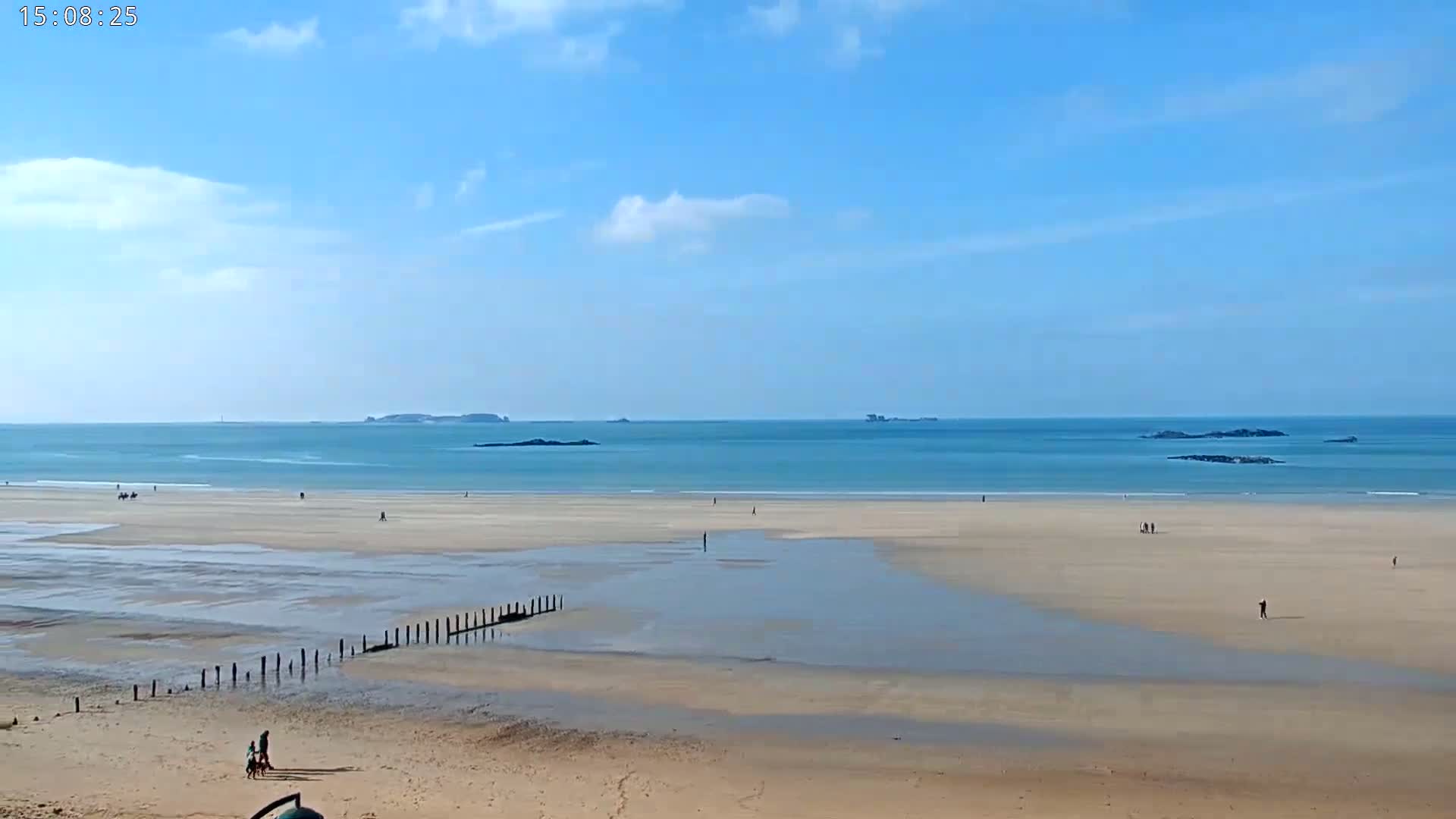 A wide sandy beach stretches to a calm, light-blue ocean under a mostly sunny sky, with a few distant islands visible on the horizon.