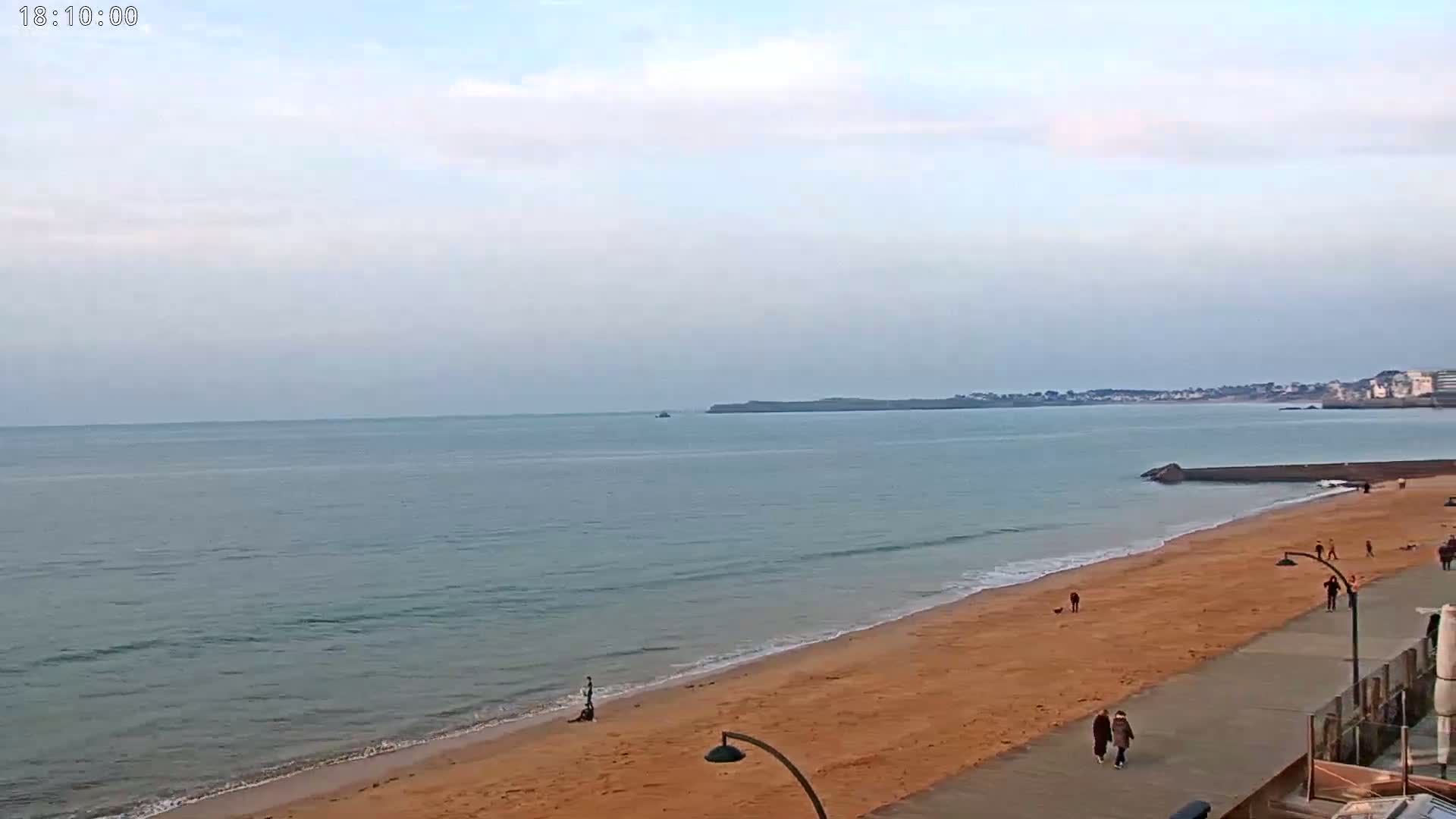 A sandy beach with a few people walking along the shore and a calm ocean under an overcast sky.