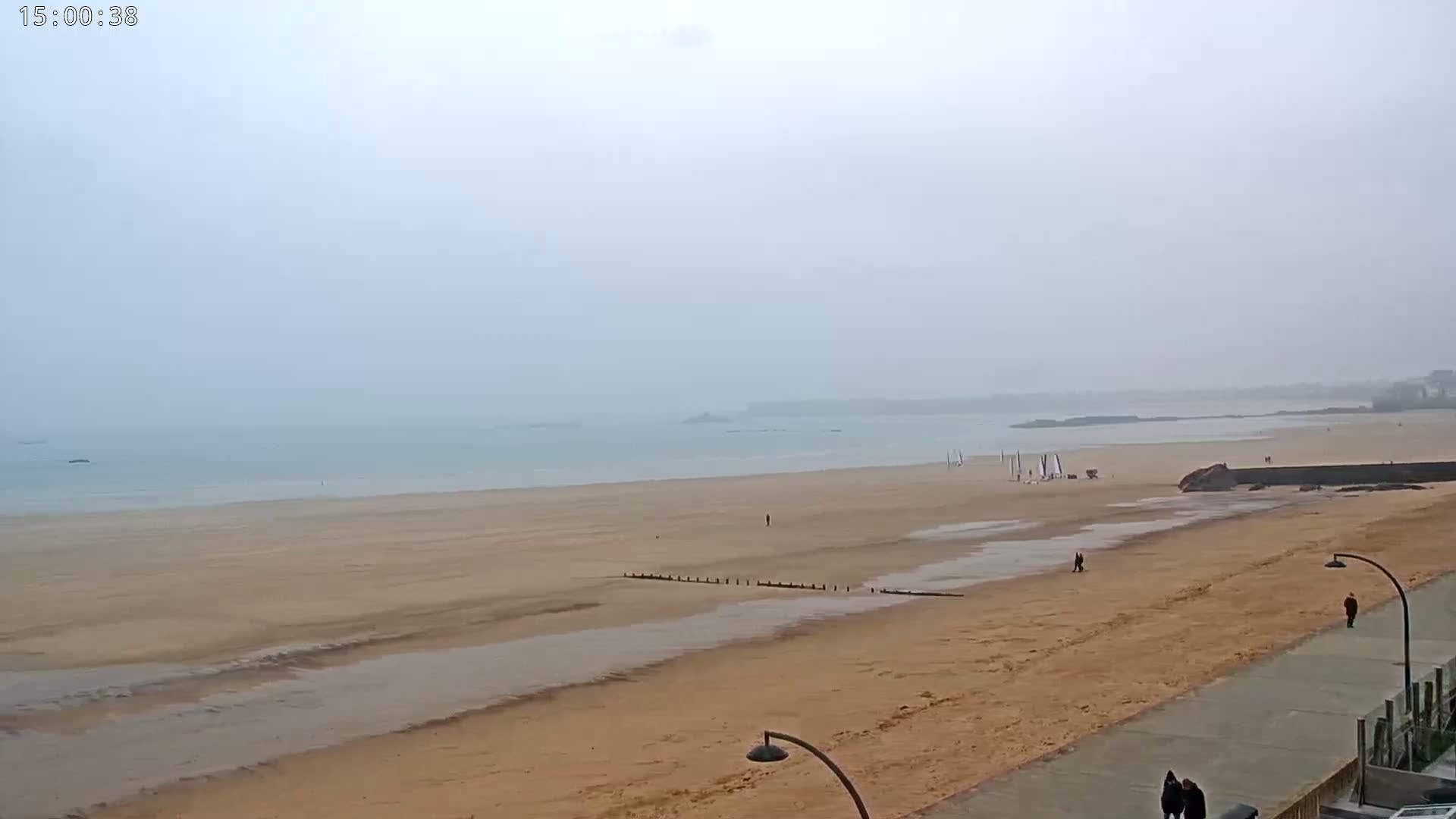 A wide, sandy beach stretches to a calm, hazy ocean under an overcast sky.
