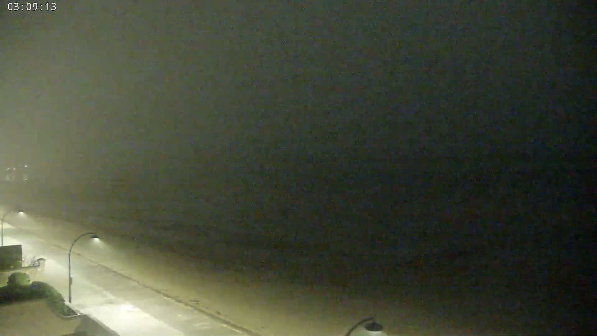 A nighttime coastal scene shows a dimly lit road next to a beach under a dark, hazy sky.