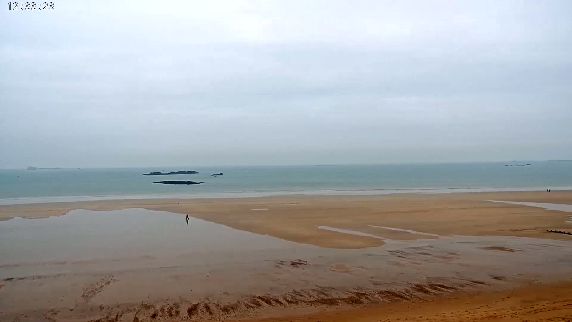 A wide sandy beach with shallow water pools stretches to a calm, grey ocean under an overcast sky, where a few small islands are visible in the distance.