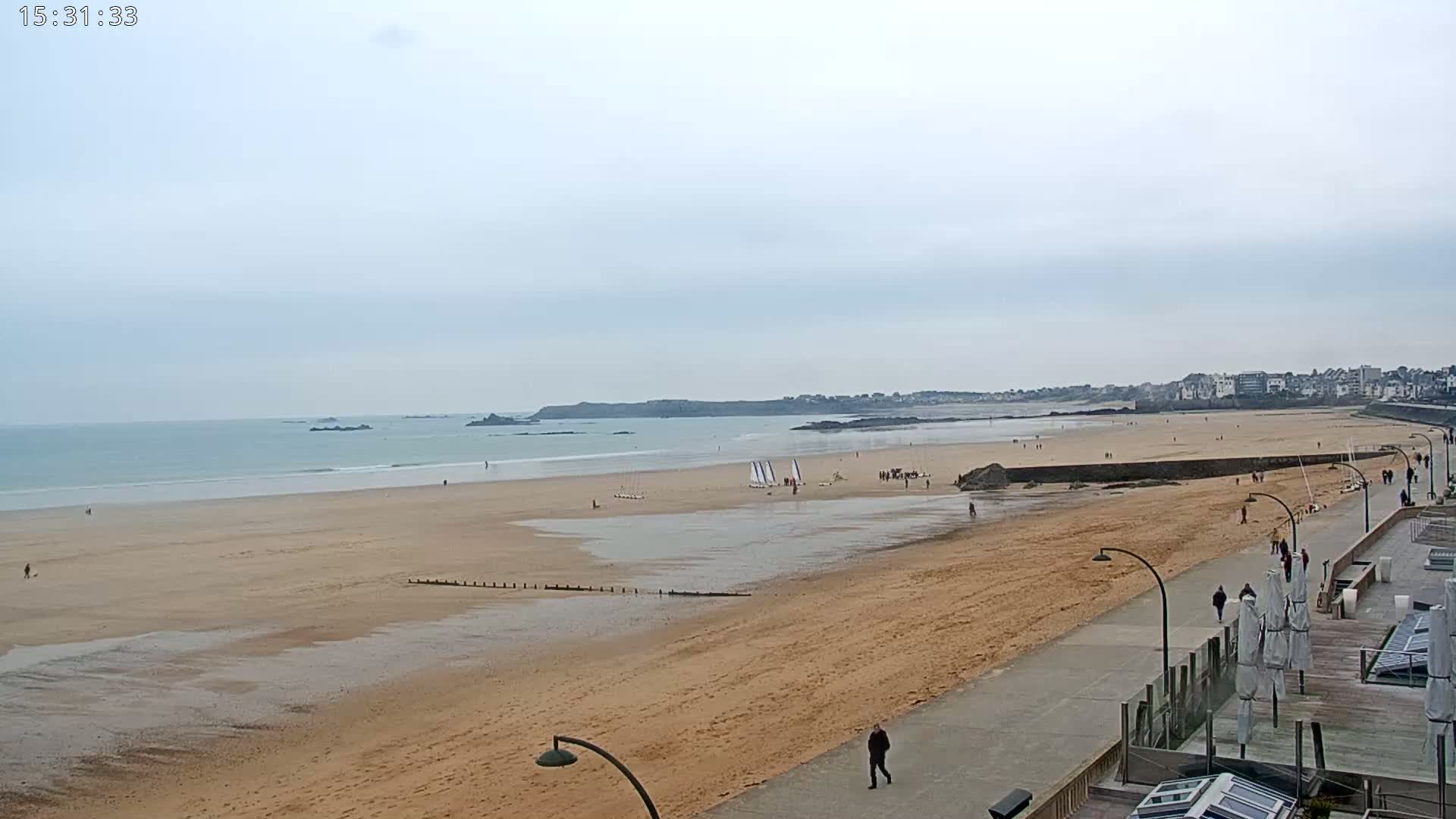 A long sandy beach stretches to a calm ocean under an overcast sky, with a few people strolling along a paved walkway and some small sailboats on the water.