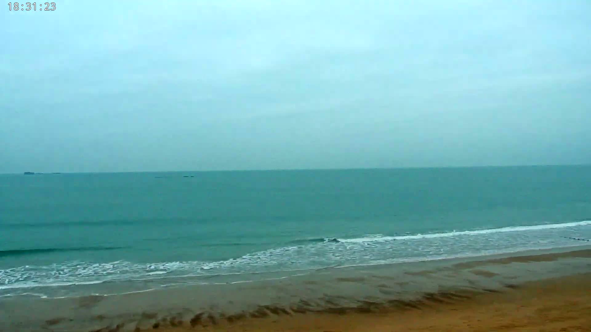 A wide sandy beach meets a calm, teal ocean under a cloudy sky.