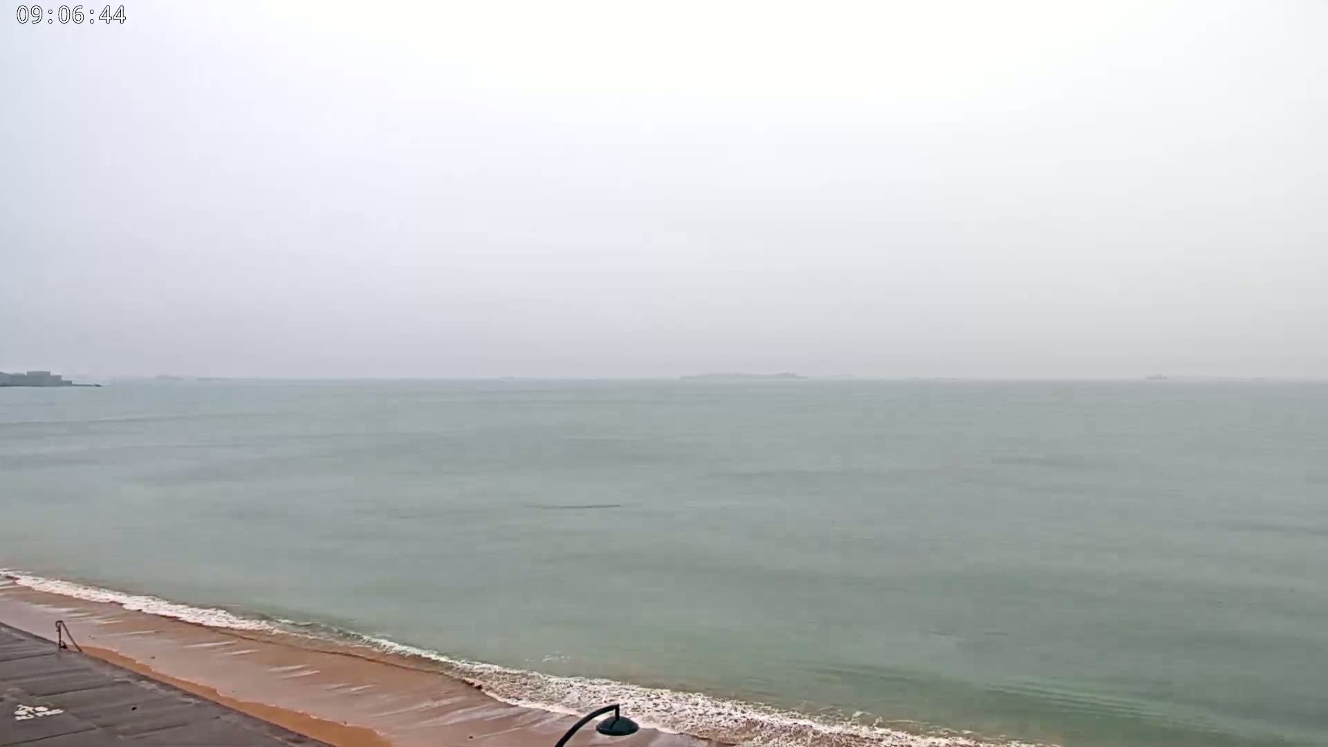 A hazy, overcast day shows a calm sea with small waves lapping a sandy beach.