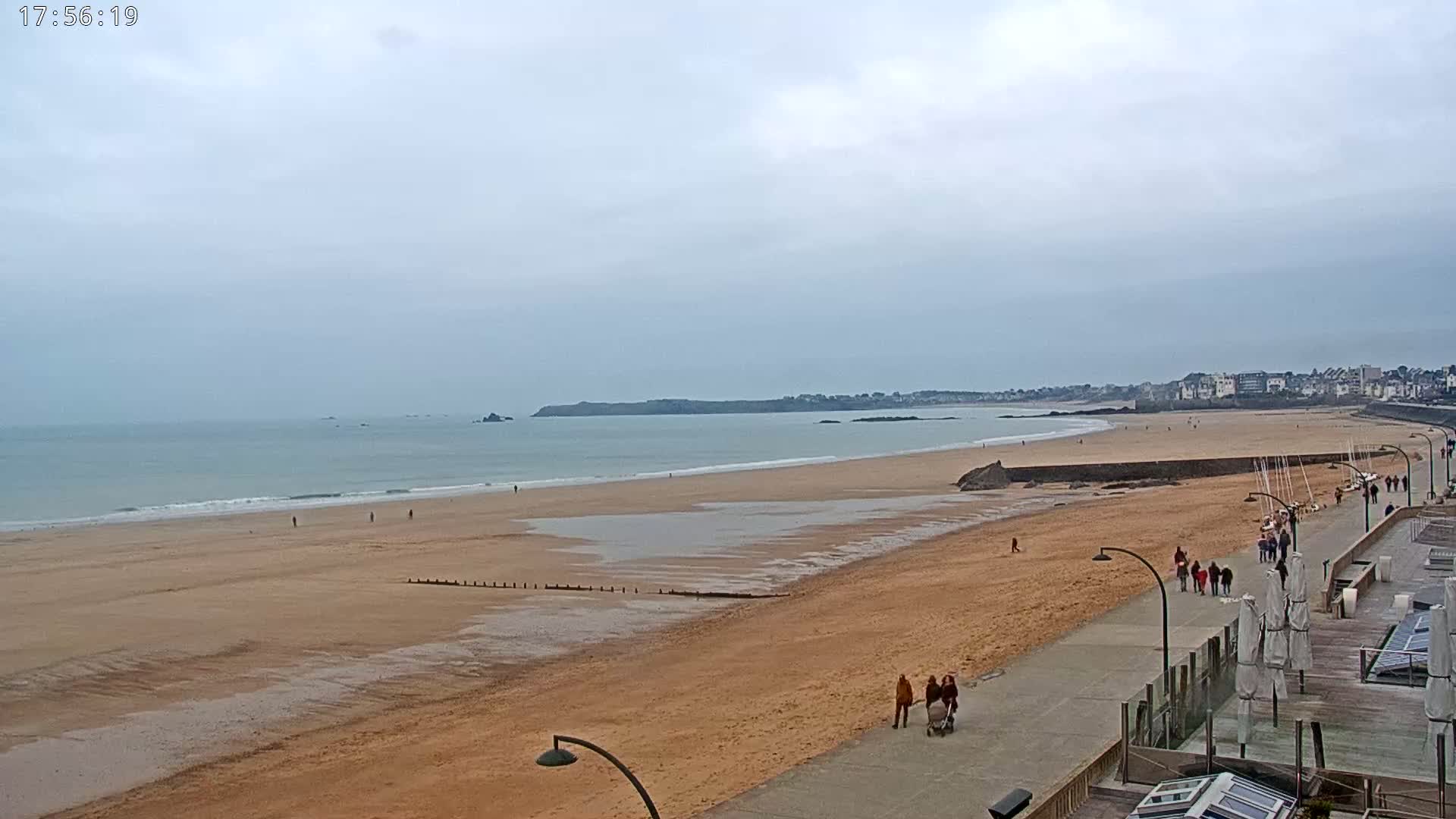 A long, sandy beach stretches out to a calm sea under a cloudy sky, with a few people walking along the shoreline and promenade.