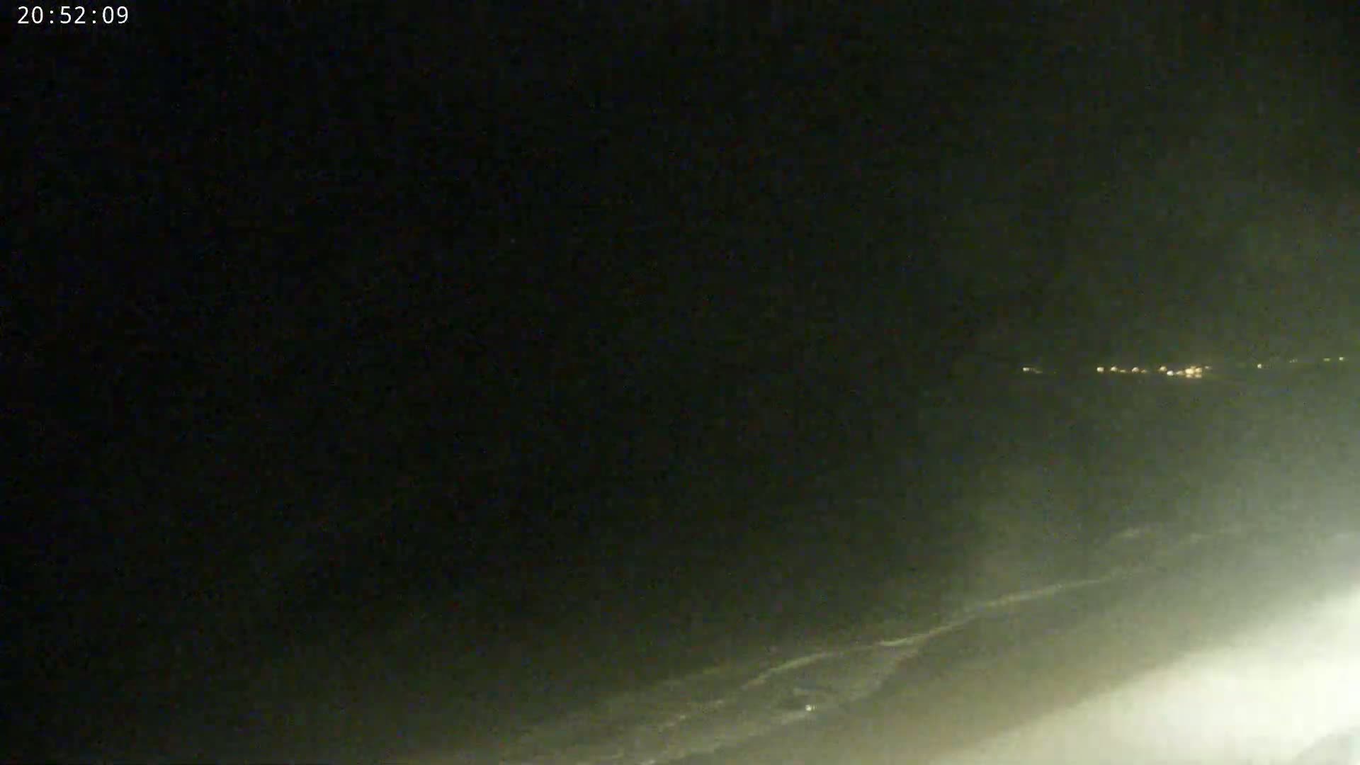A nighttime, hazy view of a coastline with dimly lit buildings in the distance and waves breaking on the shore.