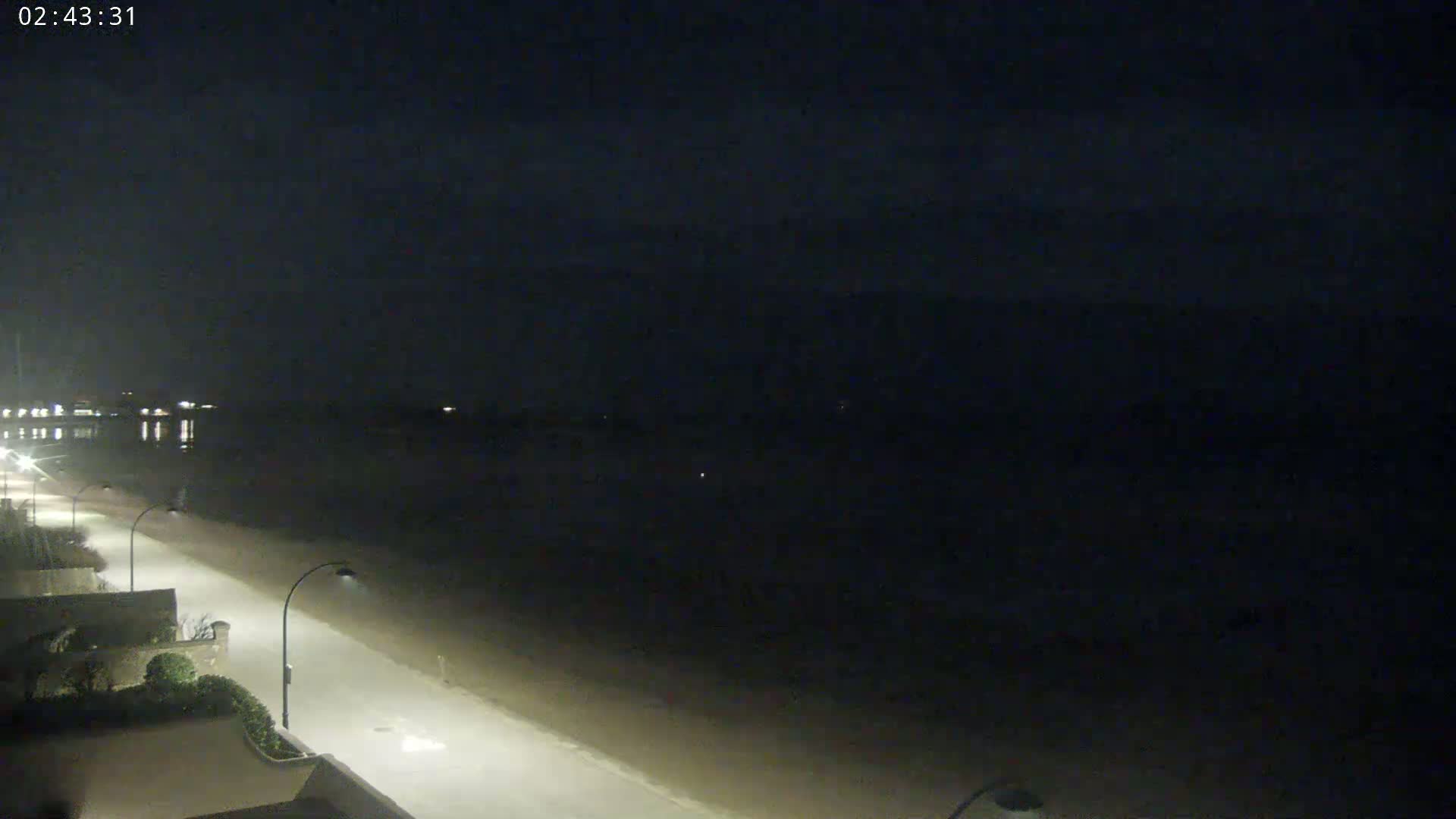 A nighttime view shows a dark, sandy beach and a dimly lit road next to it, under a cloudy sky.