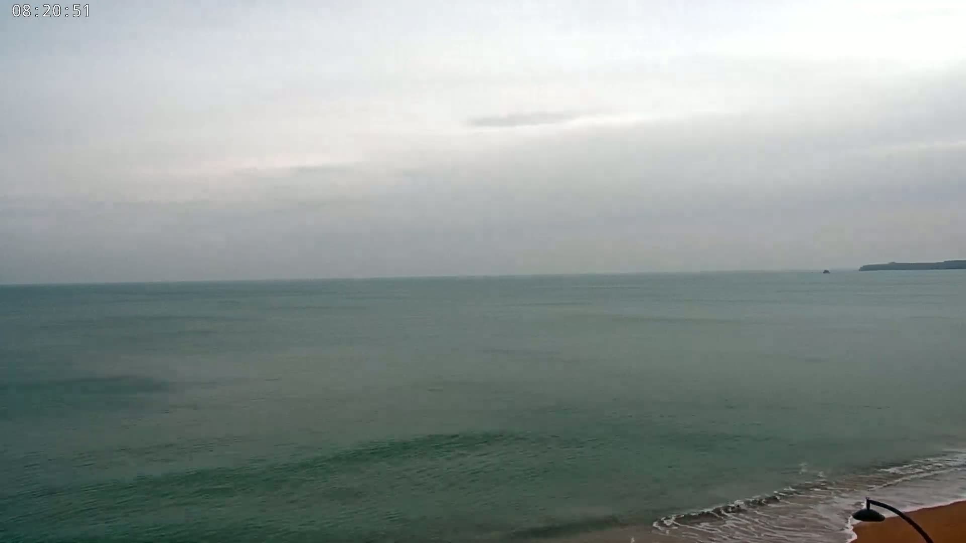 A calm, grey-skied ocean meets a sandy beach with gentle waves lapping the shore.