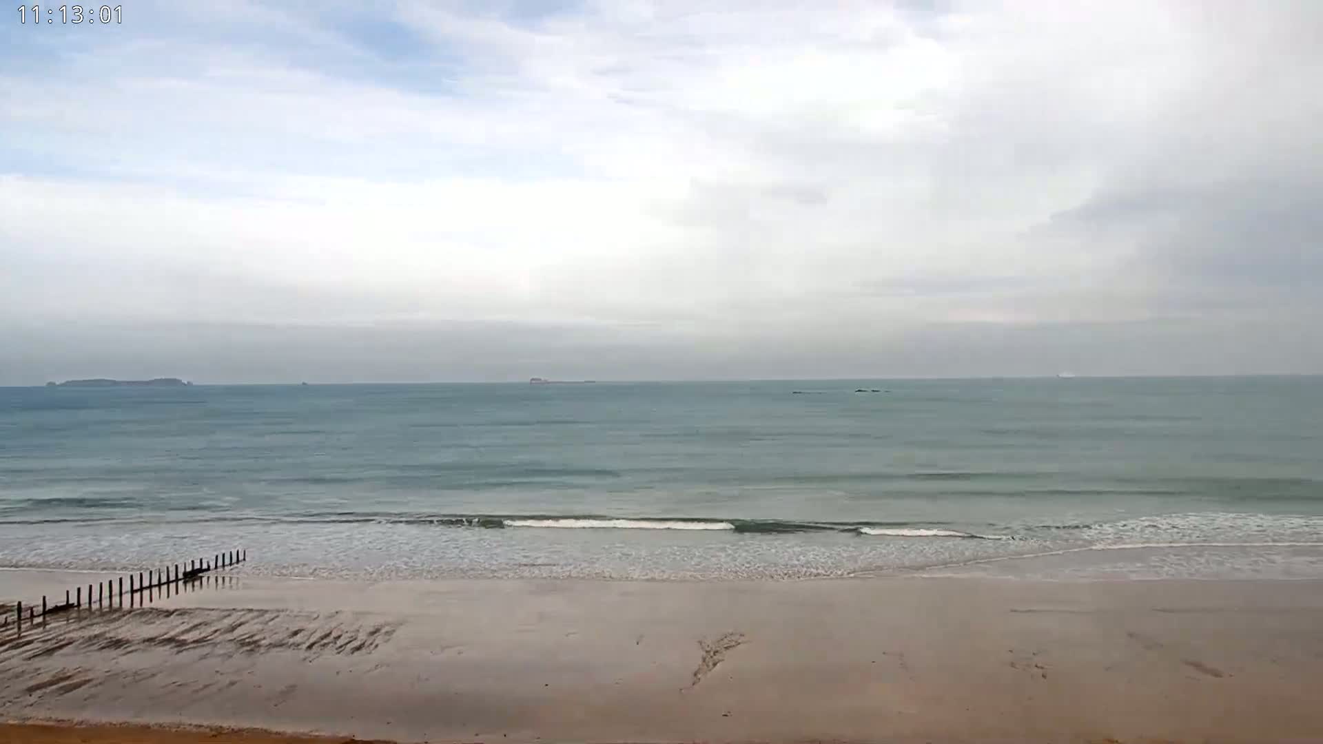 A sandy beach with small waves and a calm sea stretches towards distant islands under a mostly cloudy sky.