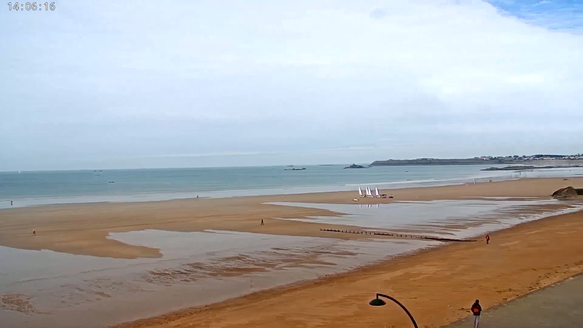 A mostly cloudy day at a wide sandy beach with a calm ocean, a few people walking, and several small sailboats in the water near the shore.