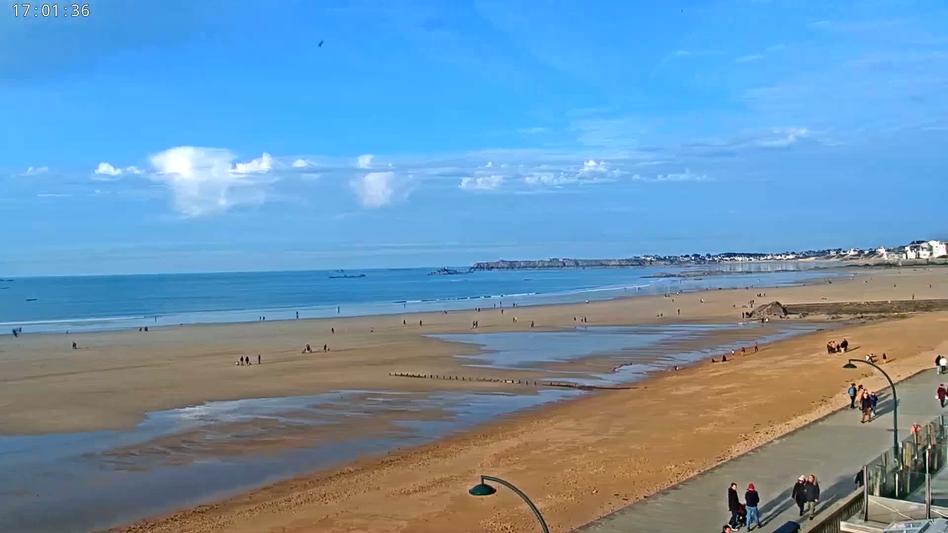 A wide sandy beach with scattered people stretches along a calm ocean under a mostly sunny sky with a few clouds.