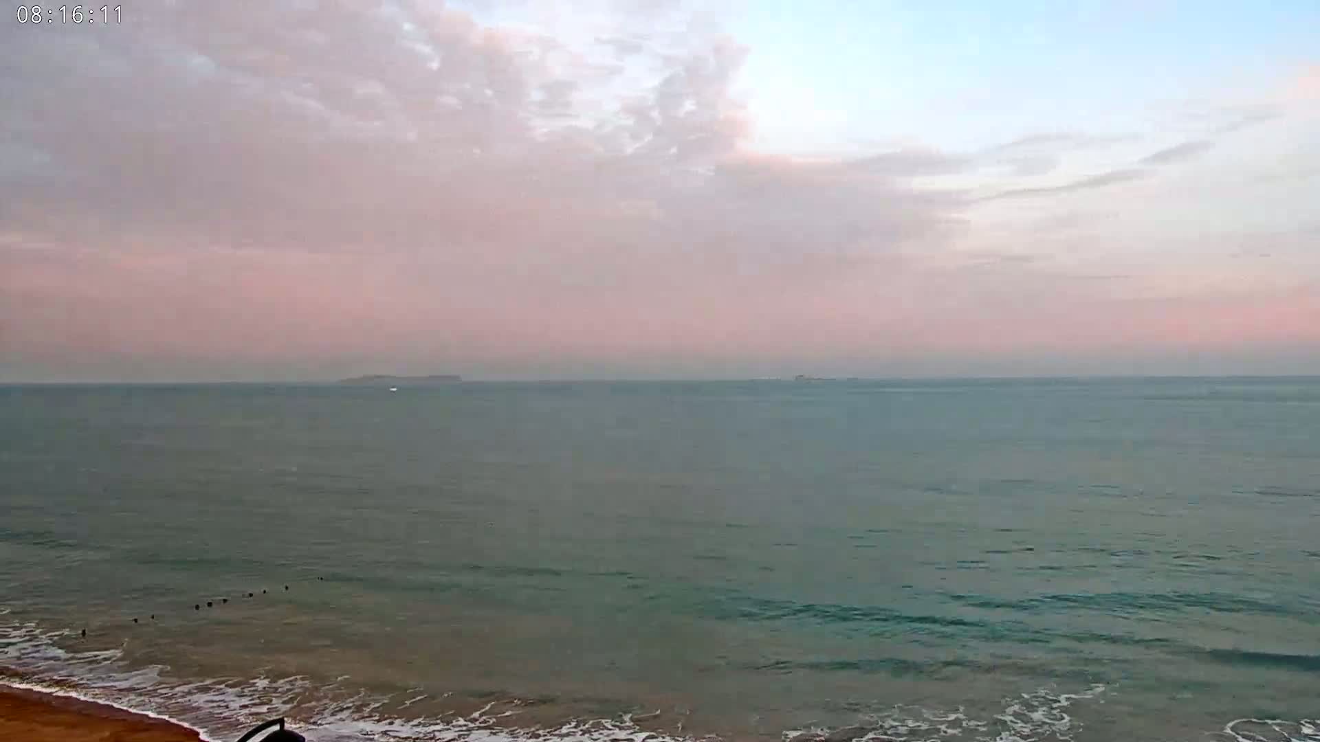 A calm ocean with a sandy beach meets a pale pink and blue sky filled with thin clouds.