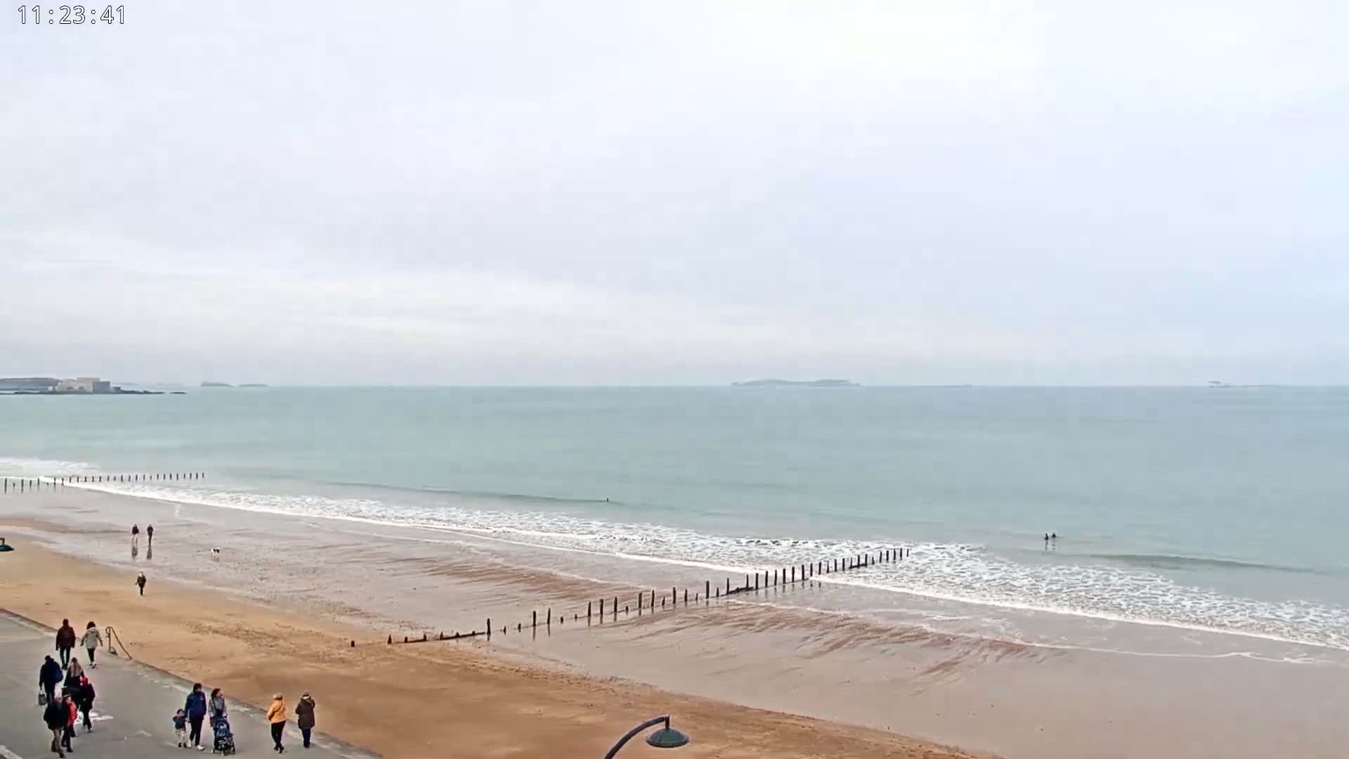 A wide sandy beach with gentle waves under an overcast sky, several people walking on the beach and boardwalk, and a line of wooden posts extending into the water.
