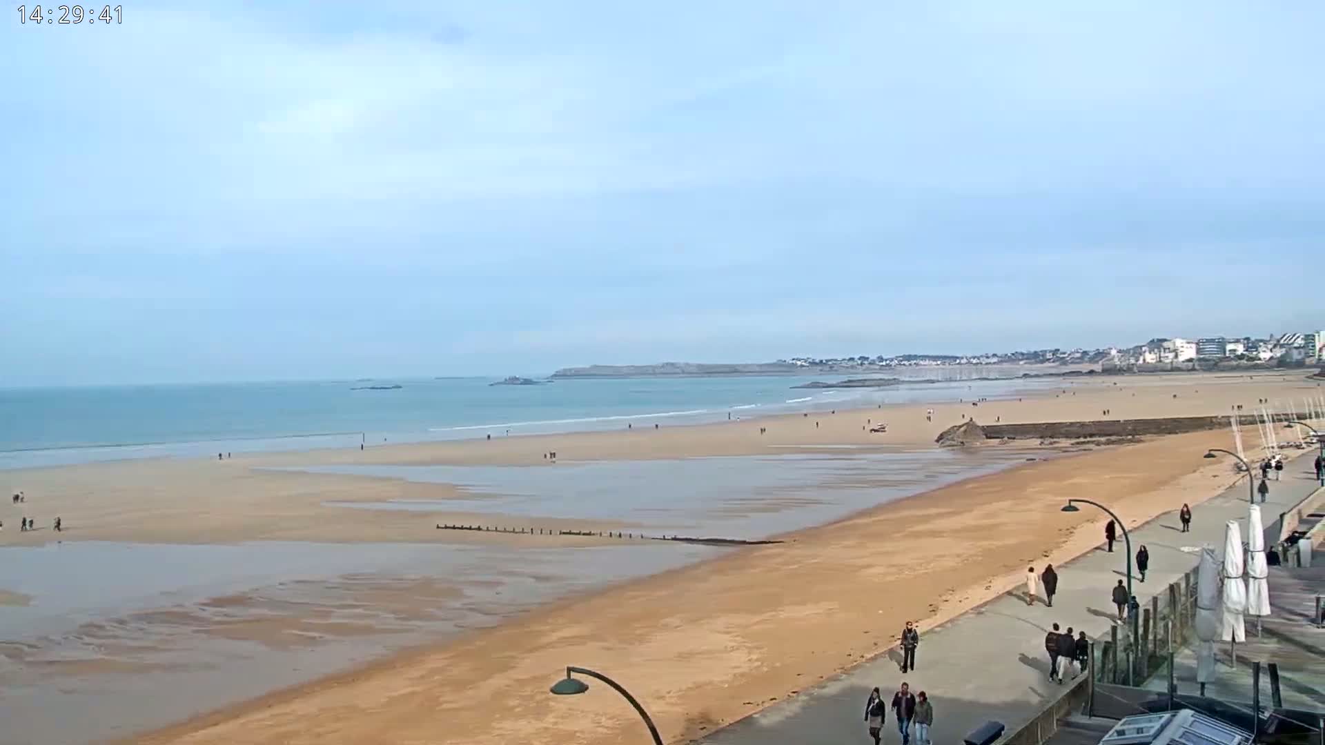 A wide sandy beach stretches along a calm ocean under a mostly clear sky, with people walking on the beach and a paved boardwalk.